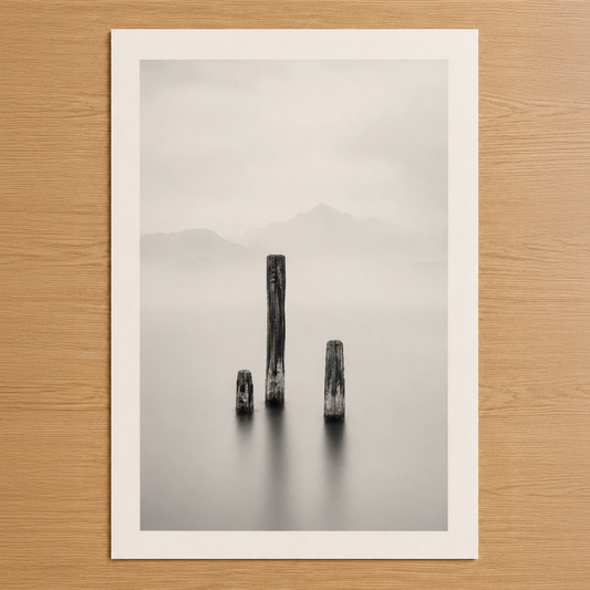 Minimalist siyah beyaz fotoğraf tablosu, sisli su içinde dikilen ağaç kütükleri ve dağ silueti gösteriyor.