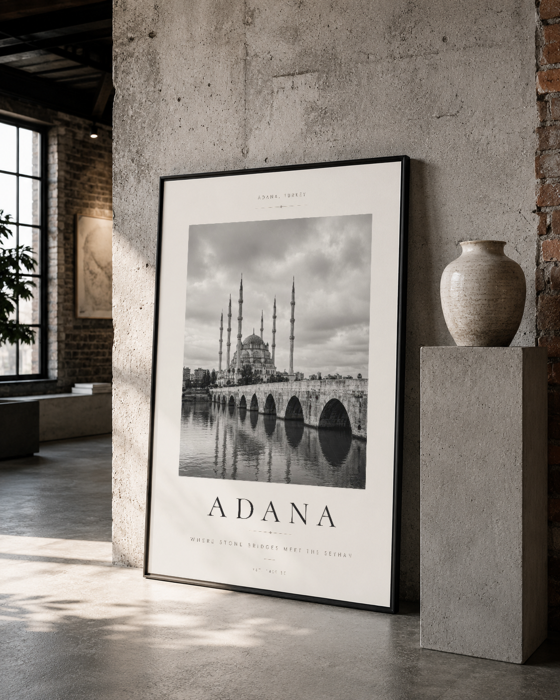 Siyah çerçeveli Adana posteri, tarihi camii ve köprü görüntüsüyle endüstriyel tarzda tasarlanmış loft ortamında duruyor.