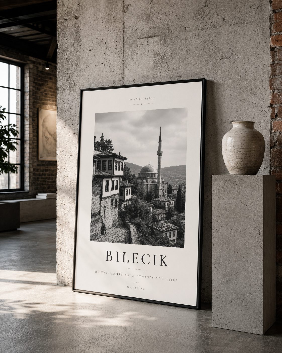 Bilecik panoramasının siyah beyaz fotoğrafı, siyah çerçevede, beton duvara asılı, yan tarafta seramik vazo ile sergilenen