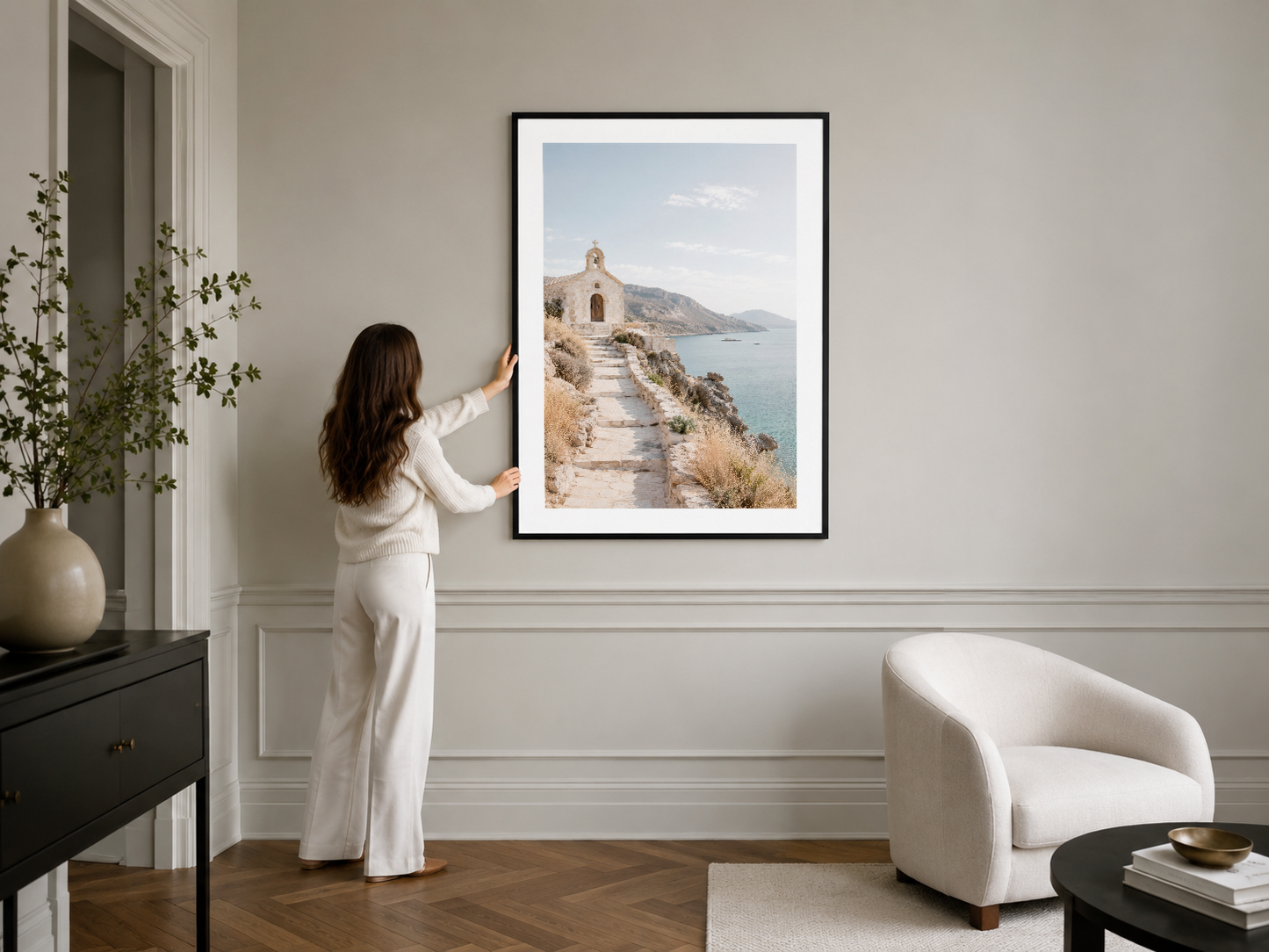 Siyah çerçeveli tablo, minimalist iç tasarımda, modern beyaz koltuğun üstünde, Akdeniz sahil manzarası sahnesi.