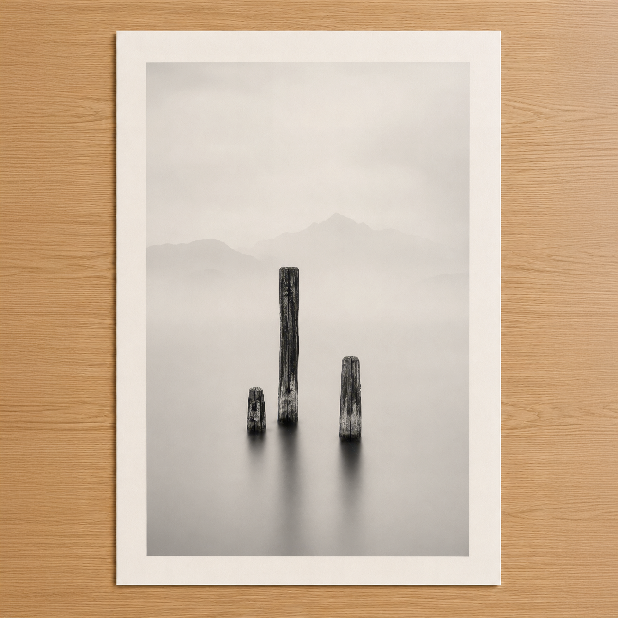 Minimalist siyah beyaz fotoğraf tablosu, sisli su içinde dikilen ağaç kütükleri ve dağ silueti gösteriyor.