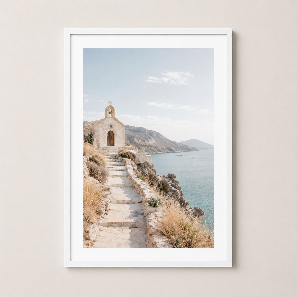 Mediterranean coastal scene with white chapel, stone pathway and mountains, white framed